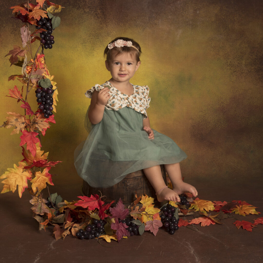 bambina con vestito verde di tulle seduta su tino con foglie autunnali e grappoli d'uva